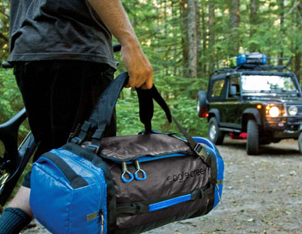 Featuring the cargo hauler duffel from Eagle Creek. A man carrying a blue and black duffel is returning to his jeep in the middle of a forest.